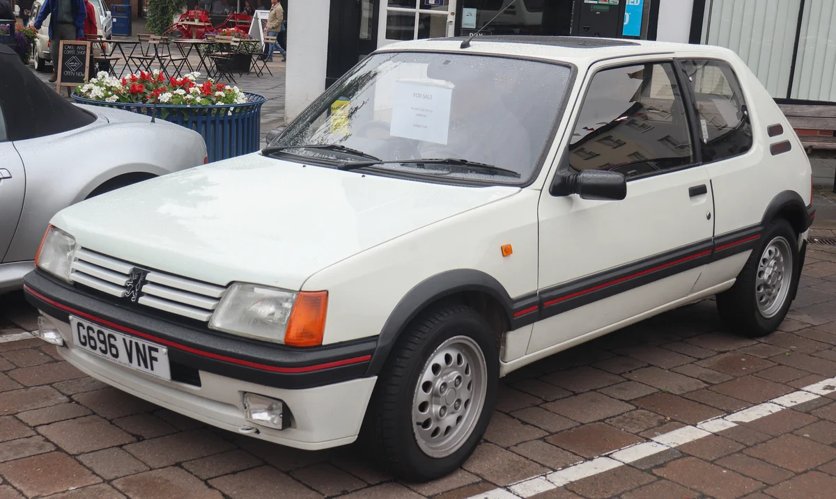 Peugeot 205 GTI 1.6 blanche de 1990 garée sur un parking pavé lors d'un rassemblement de voitures anciennes